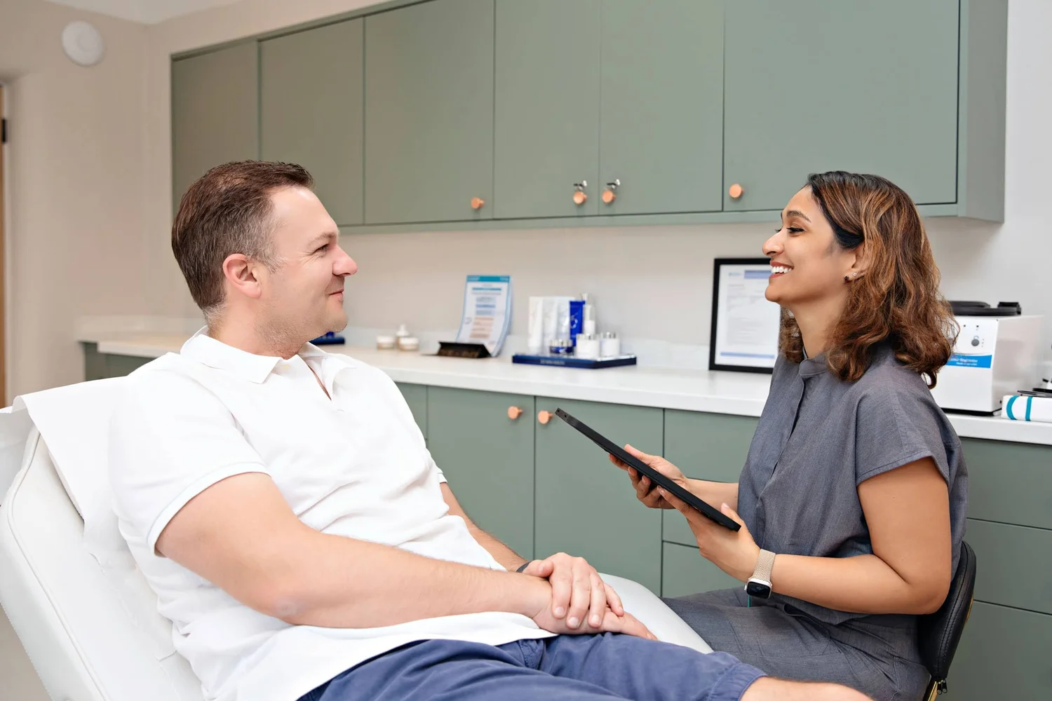 Patient relaxed in a dental chair chatting with a smiling clinician in a treatment room with green cabinets behind them.